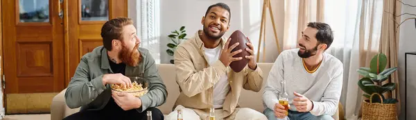 Tre uomini diversi si siedono su un divano in abbigliamento casual, abbracciando felicemente un calcio, godendo di un momento divertente e rilassato insieme. — Foto stock