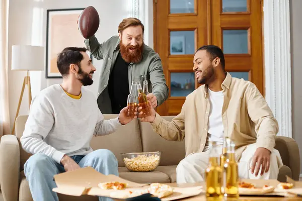 Tres hombres alegres e interraciales en atuendo casual se sientan en un sofá, uniéndose y disfrutando de la compañía de los demás. - foto de stock