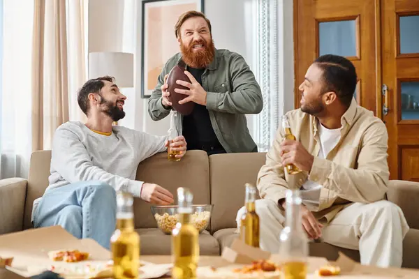 Tres hombres alegres y guapos de diferentes razas en atuendo casual, disfrutando de la compañía de los demás mientras están sentados en la parte superior de un sofá. - foto de stock