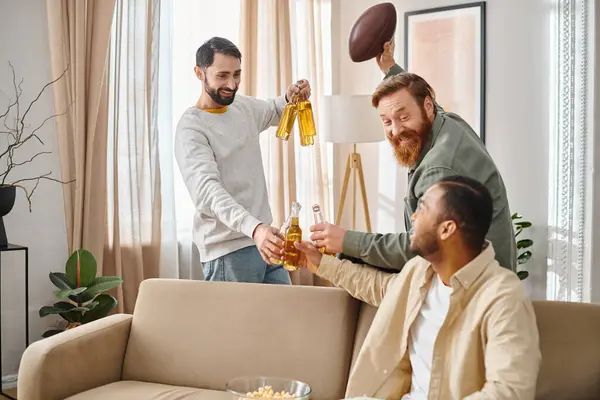 Dos hombres, uno sosteniendo una botella de cerveza, se sientan en un sofá acogedor, charlando y riendo con su atuendo casual. - foto de stock