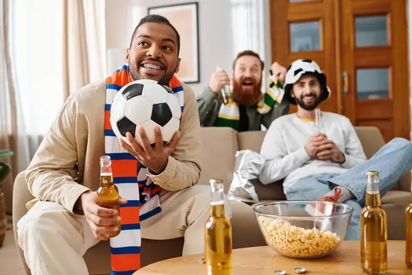 A man in casual attire holds a bottle of beer while holding a soccer ball, enjoying a cheerful moment with friends at home. — Stock Photo