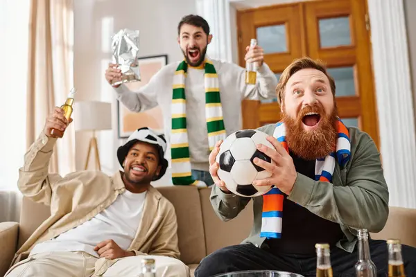 Drei fröhliche, unterschiedliche Männer in legerer Kleidung sitzen auf einer Couch und halten fröhlich Fußballbälle in der heimischen Umgebung.. — Stockfoto