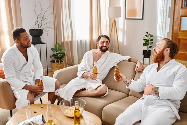 Three diverse, cheerful men wearing bathrobes have a great time chatting and laughing while sitting on top of a couch. — Stock Photo