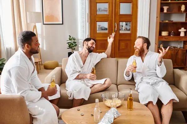 Drei bunt gemischte, gut gelaunte Männer in Bademänteln sitzen auf einer Couch und genießen in lustiger und entspannter Atmosphäre die Gesellschaft der anderen. — Stockfoto