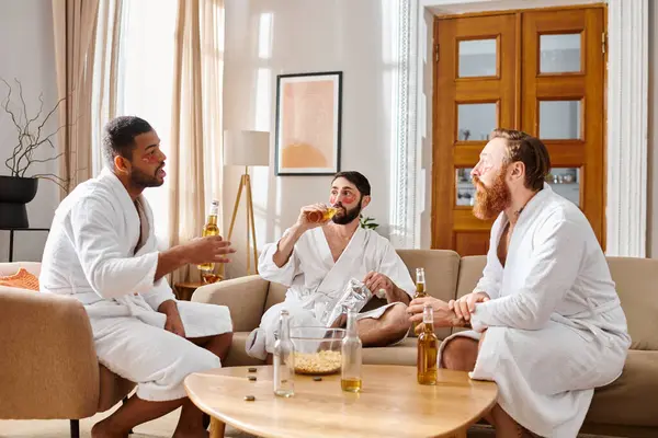 Three cheerful men of different backgrounds, in bathrobes, share laughter and camaraderie around a living room table. — Stock Photo