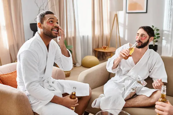 Drei unterschiedliche, gut gelaunte Männer in Bademänteln genießen eine tolle Zeit auf einer Plüschcouch und teilen Lachen und Geschichten. — Stockfoto