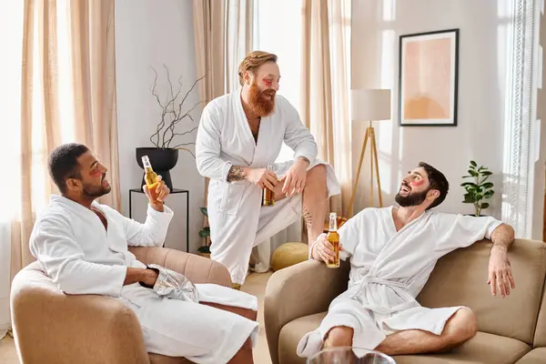 Tres hombres diversos y alegres en albornoces se sientan en un sofá, disfrutando de la compañía de los demás en un ambiente relajado. - foto de stock