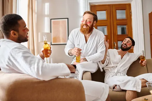 Tres hombres diversos y alegres en albornoces riendo y charlando en una acogedora sala de estar. - foto de stock
