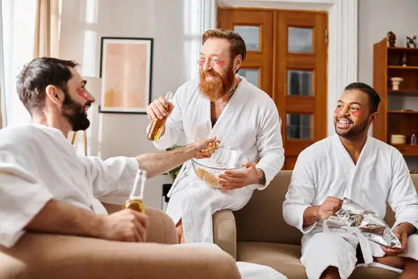 Trois hommes joyeux en peignoirs profitant d'un repas ensemble dans un cadre confortable salon. — Photo de stock
