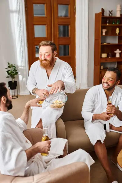 Tres hombres diversos en albornoces riendo y charlando en un acogedor ambiente de sala de estar. - foto de stock