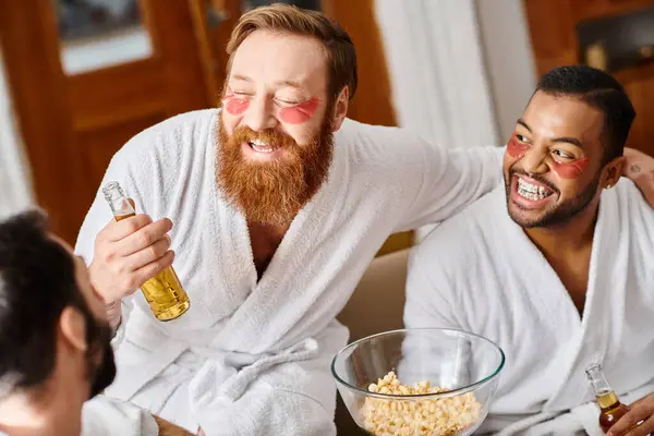 Diverse group of three cheerful men in bathrobes sitting at table, laughing and sharing a bowl of popcorn. — Stock Photo