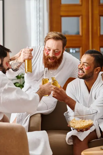 Tres hombres alegres y diversos en albornoces disfrutando del vino y la camaradería alrededor de una mesa. - foto de stock