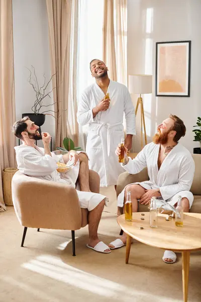 Tres hombres diversos y alegres en albornoces se sientan juntos en una sala de estar, compartiendo risas y creando recuerdos. - foto de stock