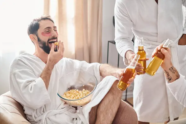 Un hombre en una silla disfrutando de un tazón de palomitas de maíz y una botella de cerveza con una sensación de relajación y satisfacción junto a sus diversos amigos. - foto de stock