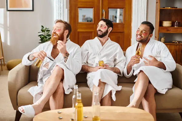 Three diverse, cheerful men wearing bathrobes are enjoying a great time together while sitting on top of a couch. — Stock Photo