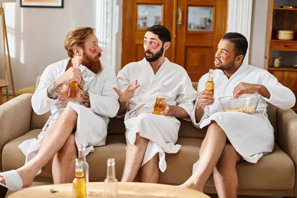Three diverse cheerful men in bathrobes enjoying each others company while sitting on top of a couch. — Stock Photo