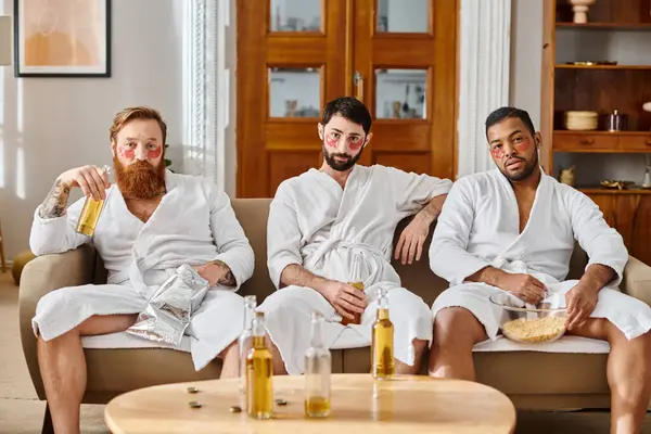 Tres hombres diversos y alegres con batas de baño, sentados encima de un sofá, disfrutando de un gran momento juntos. - foto de stock