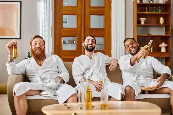 Trois hommes divers et joyeux en peignoirs assis sur un canapé, riant et tenant des bouteilles de bière. — Photo de stock