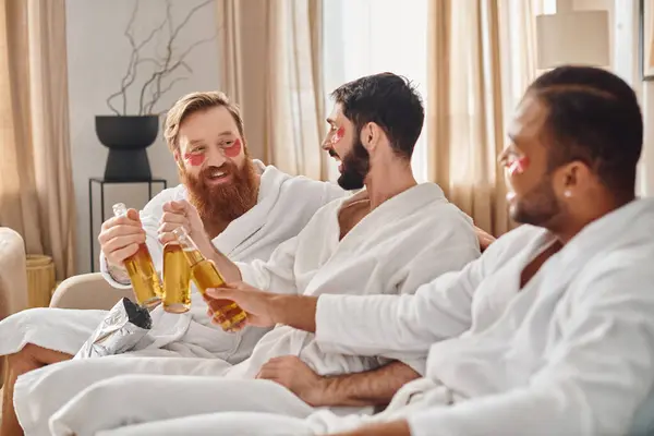 Tres hombres alegres en albornoces se sientan en un sofá, compartiendo risas y bebidas mientras disfrutan de la compañía de los demás. - foto de stock