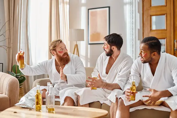 Drei bunt gemischte, gut gelaunte Männer in Bademänteln sitzen, plaudern und verbinden in gemütlicher Wohnzimmeratmosphäre. — Stockfoto