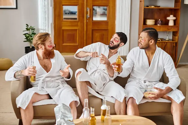 Tre uomini diversi in accappatoi si siedono sul divano, ridendo e godendosi reciprocamente la compagnia in un momento gioioso. — Foto stock