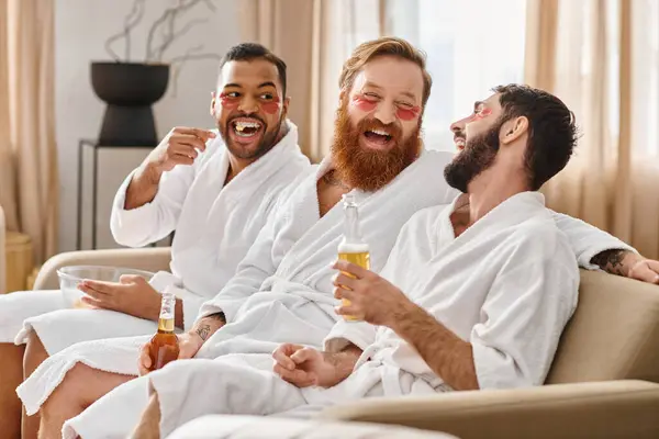 Three cheerful, diverse men in bathrobes share laughter and camaraderie while sitting on top of a plush couch. — Stock Photo