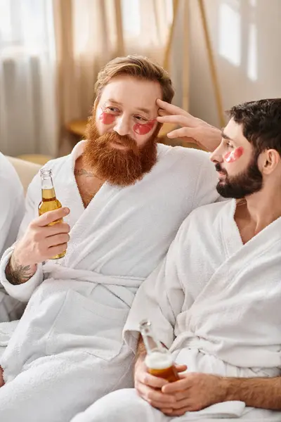 Two men in bathrobes, smiling, sit on a couch holding beer bottles, enjoying each others company and friendship. — Stock Photo