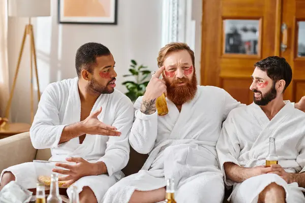 Três homens diversos e alegres em roupões de banho sentados em cima de um sofá, compartilhando risos e forjando amizades. — Fotografia de Stock