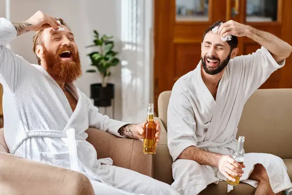 Glückliche Männer in Bademänteln lachen und plaudern, während sie in einem freudigen Moment der Freundschaft auf einer Couch hocken. — Stockfoto