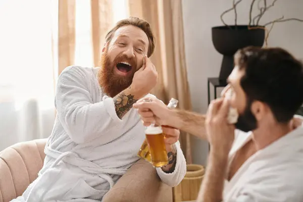 Un hombre, con una bata de baño, se relaja mientras sostiene alegremente una cerveza junto a su amigo en una alegre reunión. - foto de stock