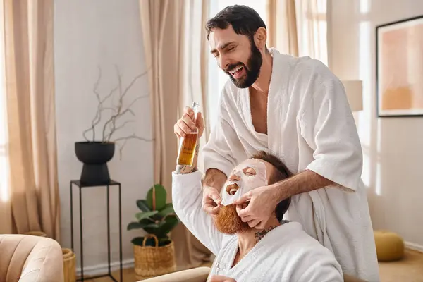 Un hombre se relaja mientras su amigo se aplica una máscara facial, parte de una experiencia de spa compartida por amigos felices en batas de baño. - foto de stock