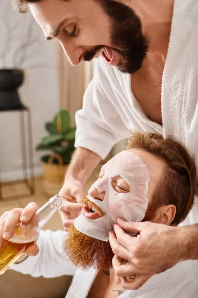 Dos hombres alegres en albornoces disfrutan de un momento de unión mientras uno coloca suavemente la máscara en la cara de otro hombre. - foto de stock