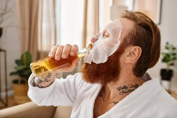 Стильный мужчина с бородой и белым халатом изящно потягивает из стакана, олицетворяя изысканность и расслабление. — стоковое фото