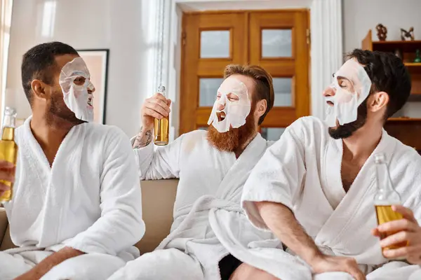 Tres hombres diversos y alegres en batas de baño y máscaras faciales disfrutando de la compañía de los demás en un sofá acogedor. - foto de stock