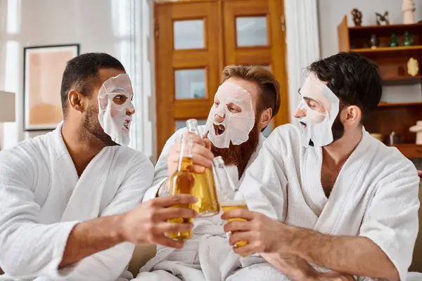 Tres hombres diversos en batas de baño, máscaras, disfrutando de cervezas juntos. - foto de stock