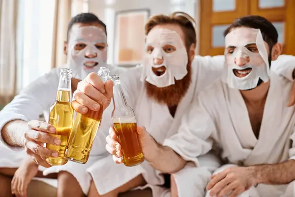 Tres hombres diversos y alegres en albornoces, con máscaras faciales, disfrutan de un momento divertido juntos, tintineando vasos de cerveza. - foto de stock