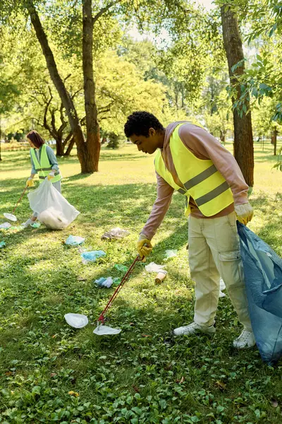 Hombre afroamericano en un chaleco amarillo de seguridad limpia la hierba en un parque, trabajando junto a su esposa, pareja socialmente activa en guantes. - foto de stock