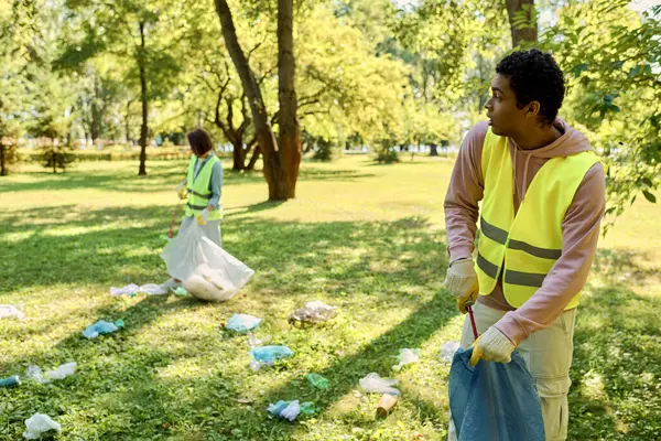 Diversi, socialmente attiva coppia indossa giubbotti di sicurezza e guanti stare nell'erba lussureggiante, la pulizia del parco insieme. — Foto stock