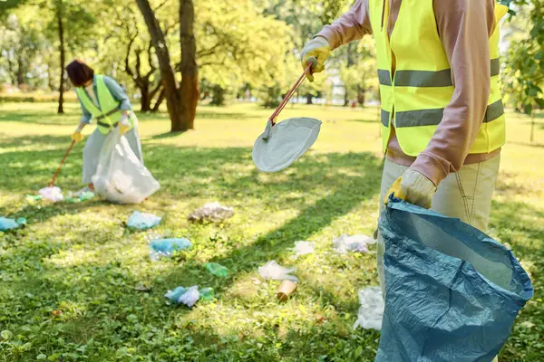 A diverse couple, wearing safety vests and gloves, cheerfully cleaning a park together, surrounded by lush green grass. — Stock Photo