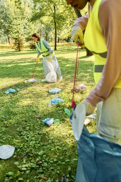 A socially active, diverse loving couple in safety vests and gloves clean a park as a group stands on a lush green field. — Stock Photo