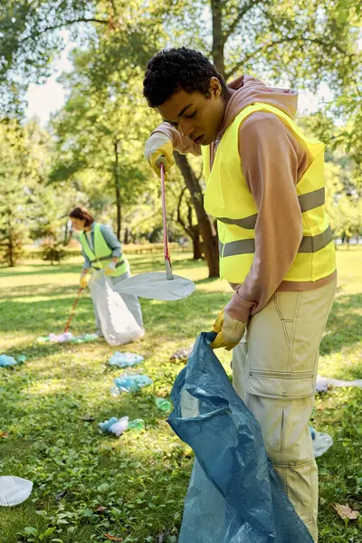 Diversa pareja en un chaleco amarillo está sosteniendo una bolsa azul mientras participa en una limpieza del parque con una pareja amorosa diversa socialmente activa. - foto de stock