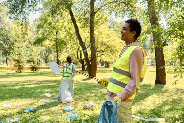Una pareja cariñosa y diversa en chalecos de seguridad y guantes limpian un parque juntos, de pie en la exuberante hierba verde. - foto de stock