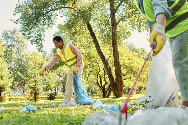 Una pareja diversa, con chalecos y guantes de seguridad, trabajando juntos para limpiar un parque cubierto de hierba. - foto de stock