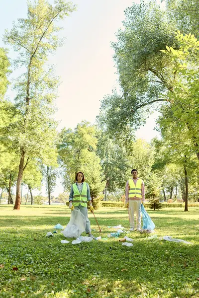 Un couple aimant, socialement actif et diversifié dans des gilets de sécurité et des gants debout dans l'herbe, nettoyant le parc ensemble. — Photo de stock