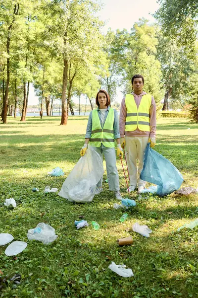 A socially active diverse couple in safety vests and gloves standing together in the lush green grass, cleaning the park. — Stock Photo