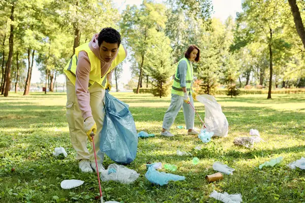 Diversa pareja con chalecos de seguridad y guantes de limpieza parque, de pie en la hierba verde exuberante con unidad y amor. - foto de stock