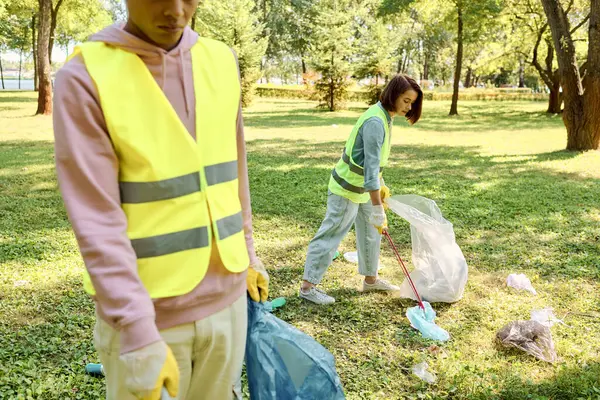 Una pareja socialmente activa y diversa en chalecos y guantes de seguridad se encuentra en la exuberante hierba verde, limpiando el parque juntos. - foto de stock