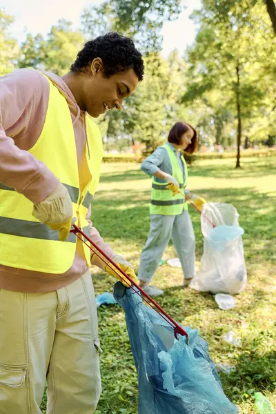 Coppia socialmente attiva e diversificata in giubbotti di sicurezza e guanti che puliscono il parco insieme ai sacchi della spazzatura. — Foto stock