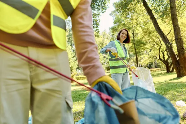 Una mujer con un chaleco de seguridad amarillo sostiene una bolsa azul mientras se ofrece como voluntaria en una limpieza del parque con su pareja. - foto de stock