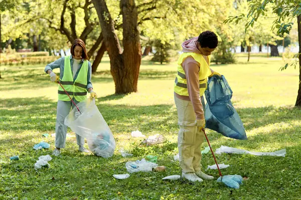 Um casal diversificado e amoroso vestindo coletes de segurança e luvas em pé na grama, limpando o parque juntamente com cuidado e unidade. — Fotografia de Stock